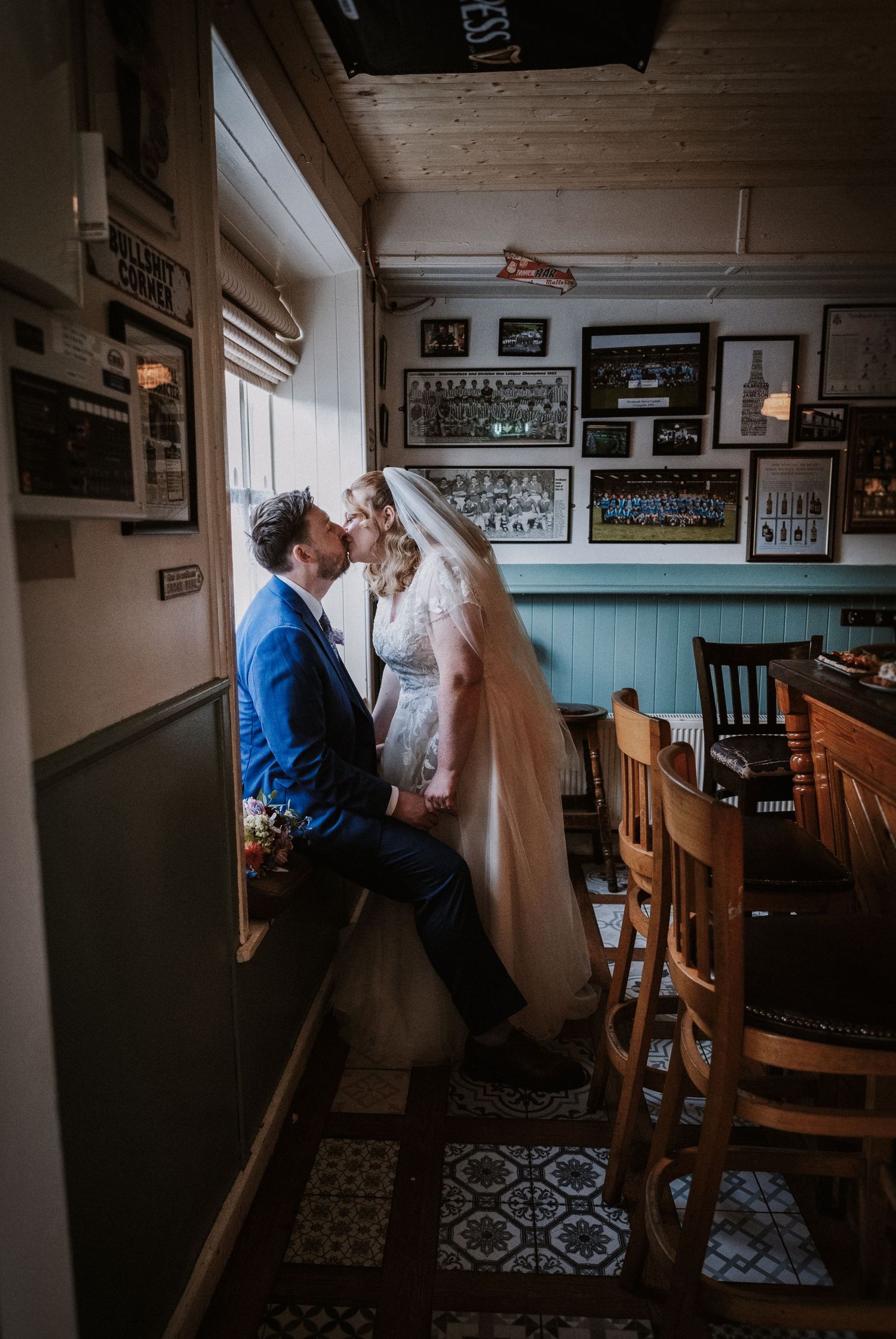 Bride and groom sharing a kiss while seated by a window inside a rustic pub venue.