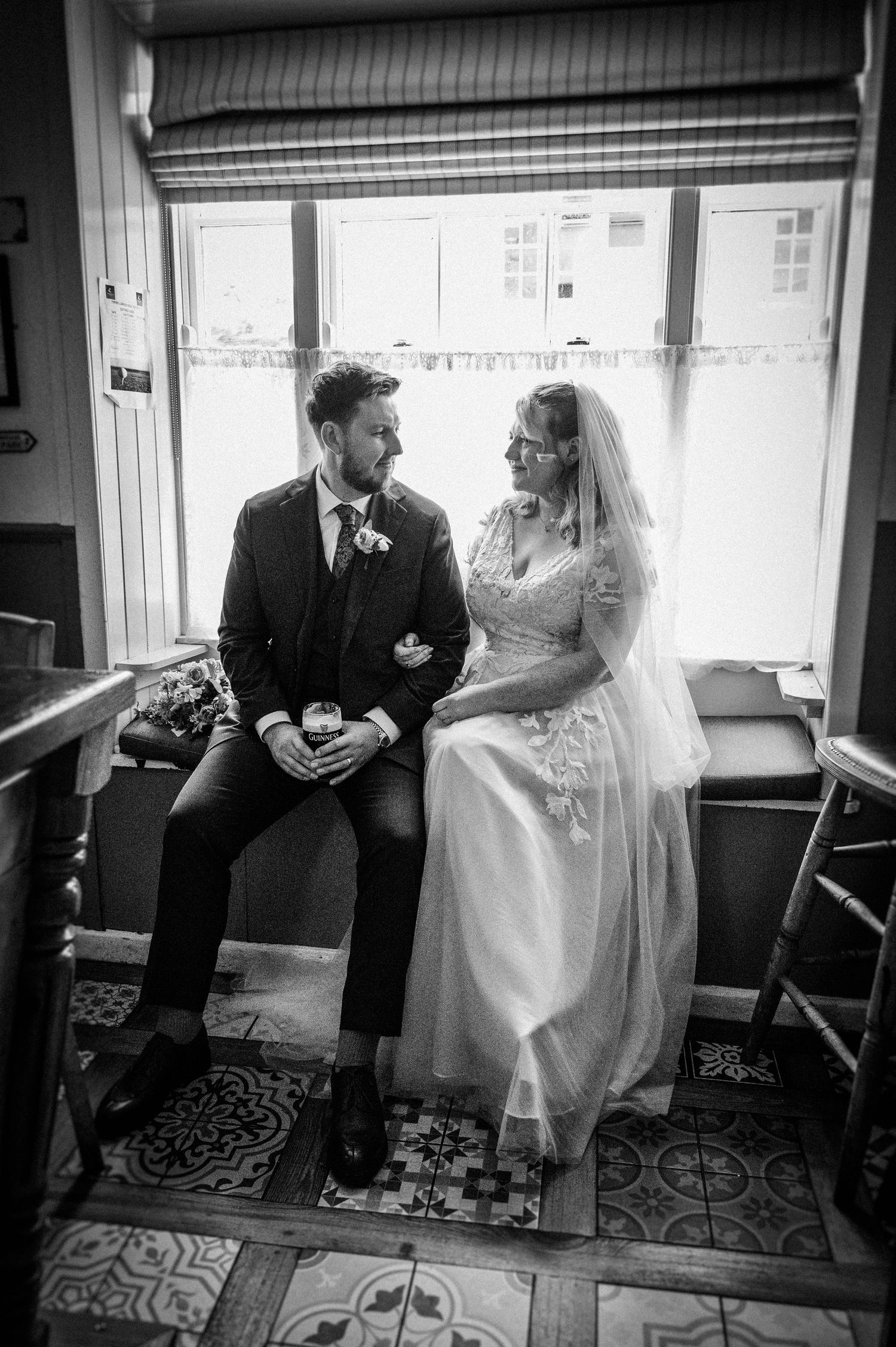 Black and white photo of a bride and groom sitting together by a window, smiling.