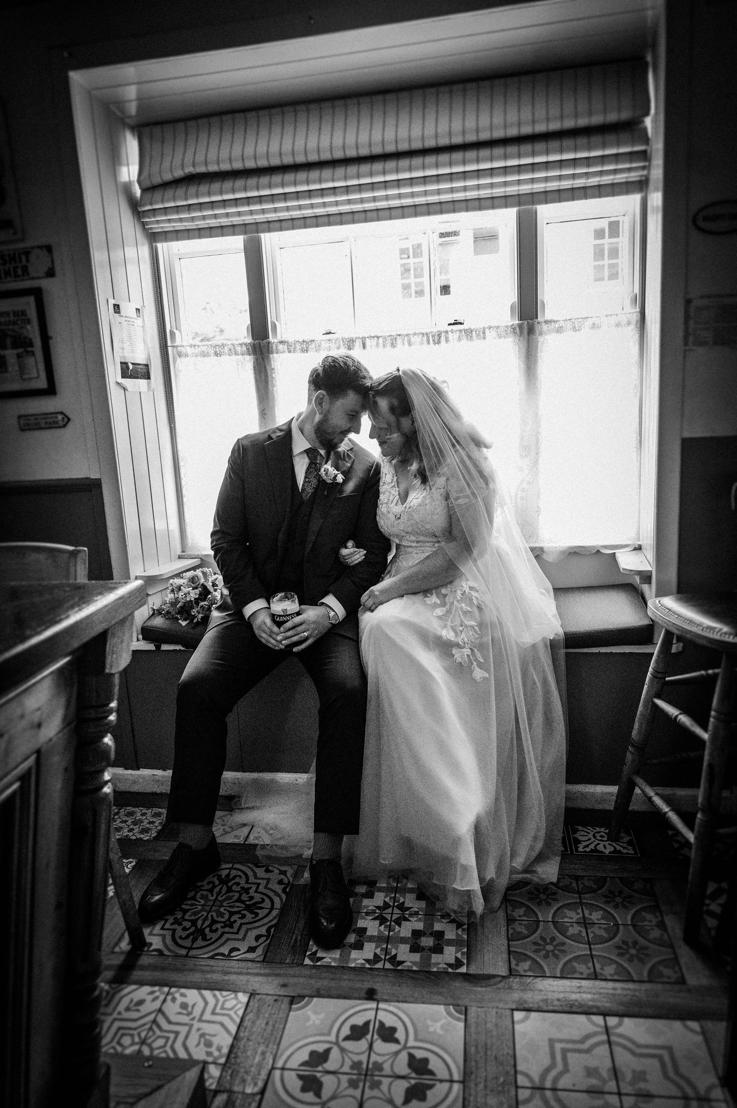 Bride and groom sharing intimate moment seated by window in black and white wedding portrait.