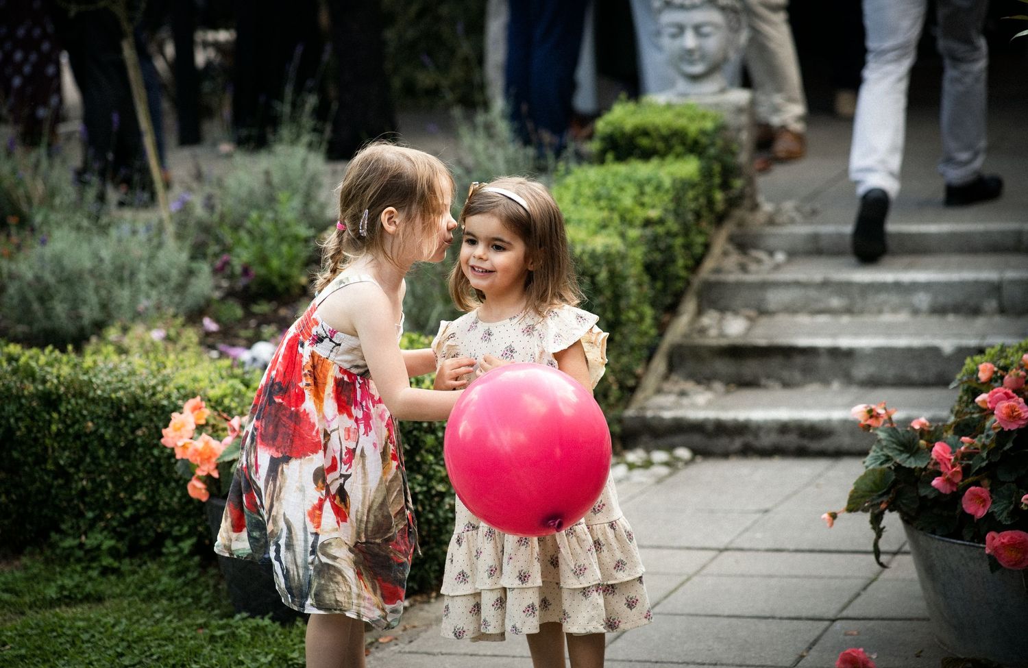 Two young girls in floral dresses playing with a pink balloon in a garden.