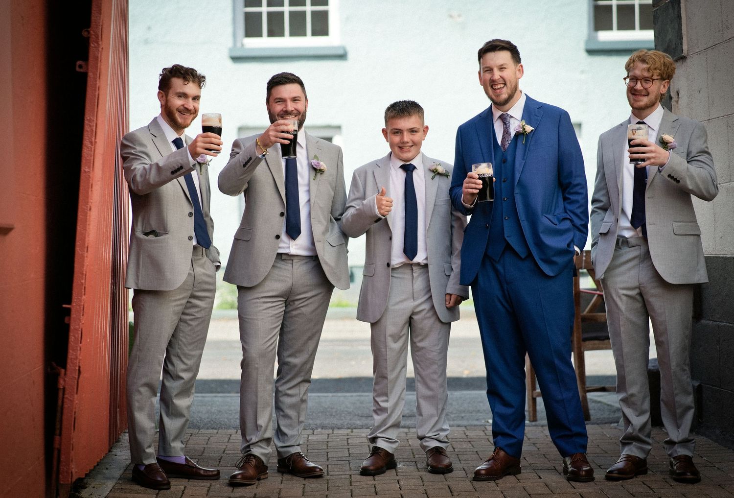 Five groomsmen in grey and blue suits smiling and holding drinks outdoors at a wedding celebration.