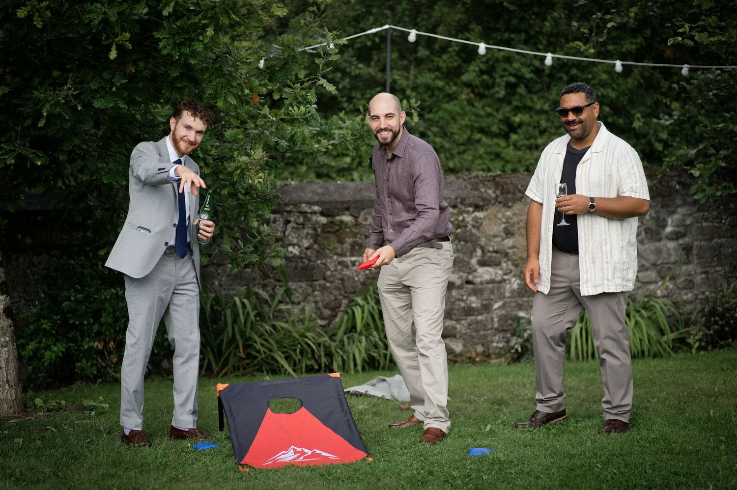 Three male wedding guests smile while playing cornhole lawn game in lush garden setting.