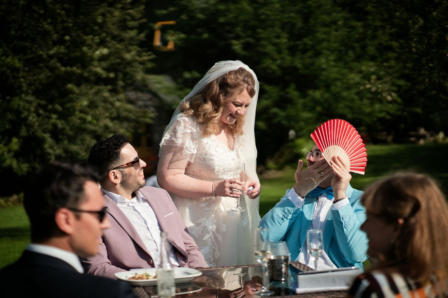 Bride in veil chatting with wedding guests seated outdoors at reception table in sunny garden.