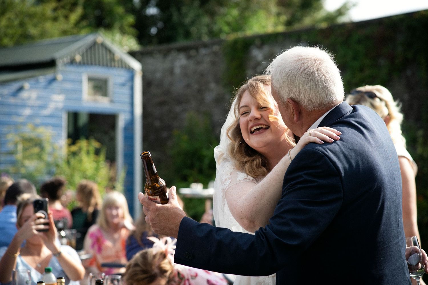 Bride laughing joyfully while dancing with older man holding beer bottle at outdoor wedding reception.