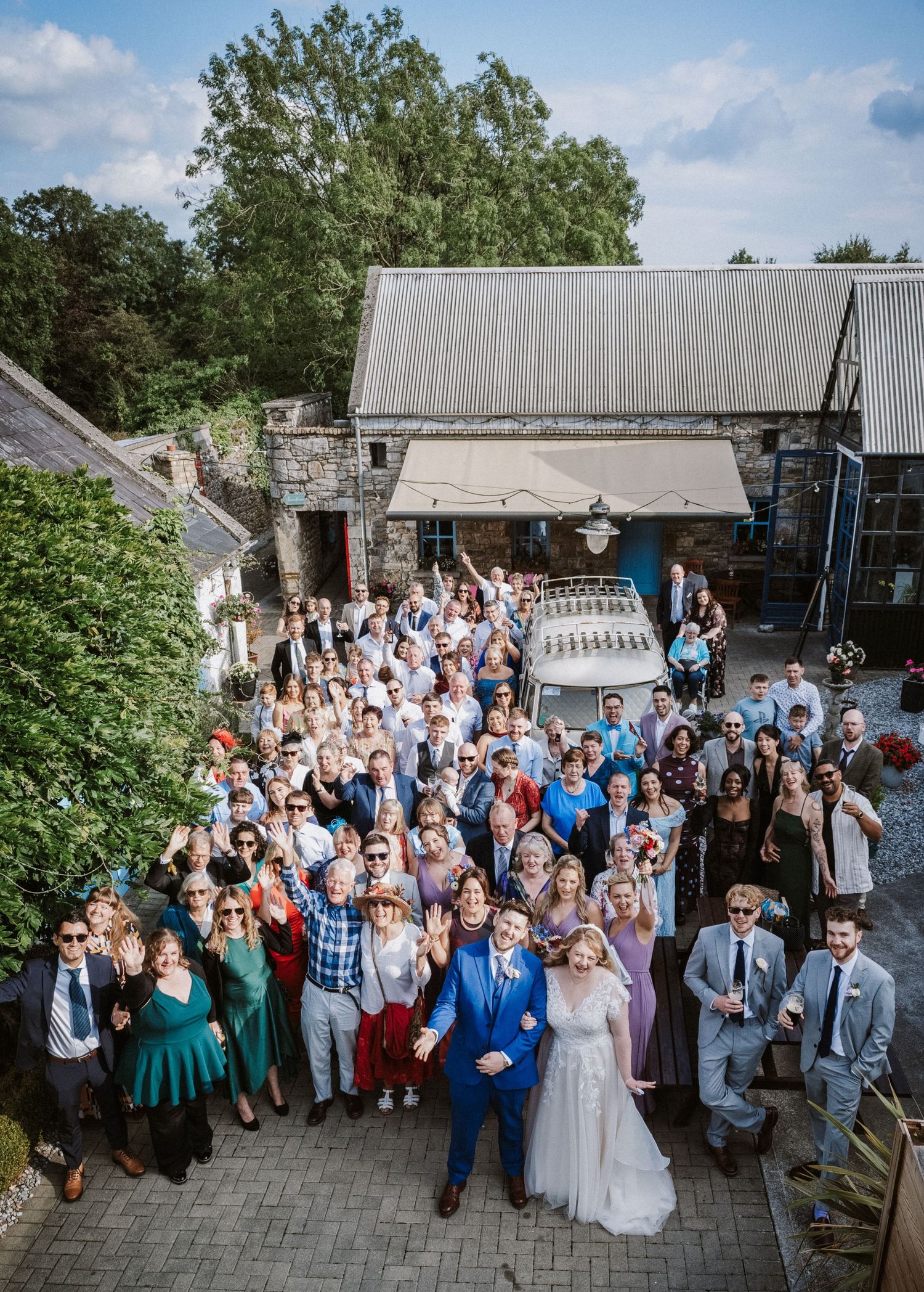 Aerial group photo of wedding party and guests outside rustic stone venue.