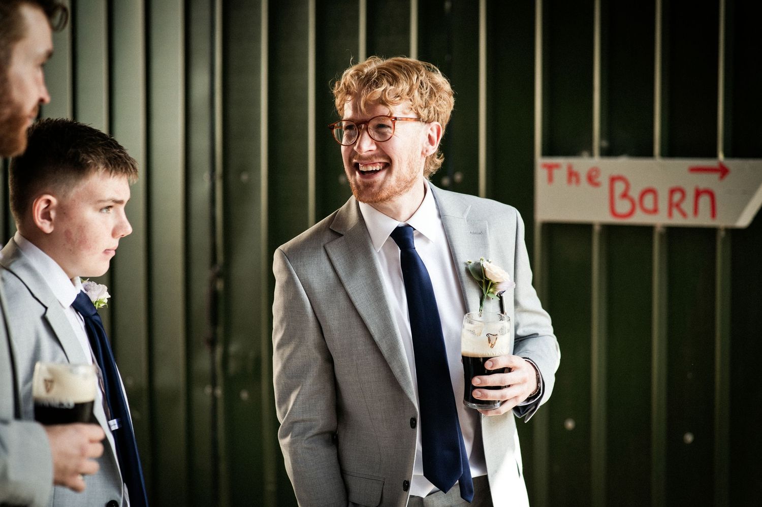 Groom laughing with groomsmen in grey suits and navy ties outside a rustic barn wedding venue.