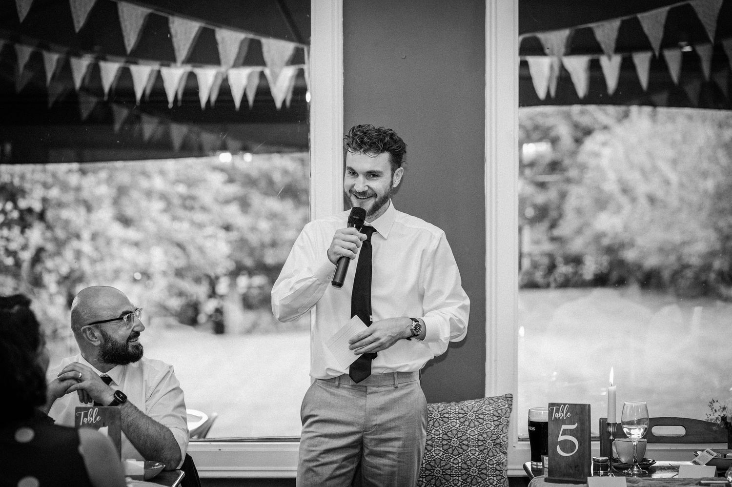 Best man delivers a heartfelt speech at a wedding reception with bunting in background.