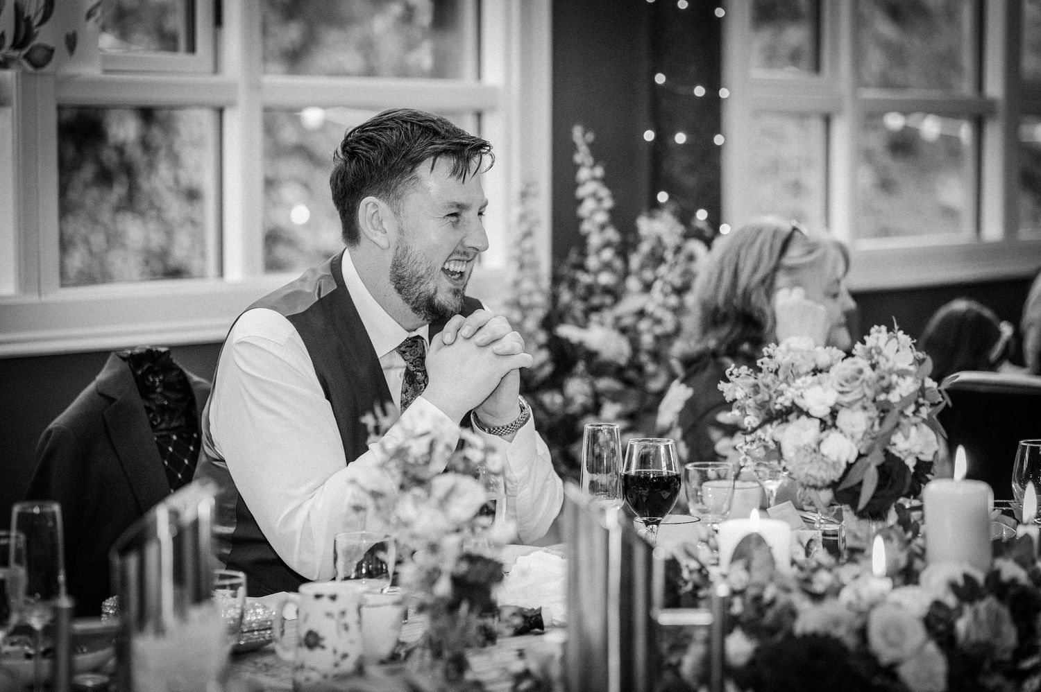 Black and white photo of a groom laughing at the wedding reception dinner table.