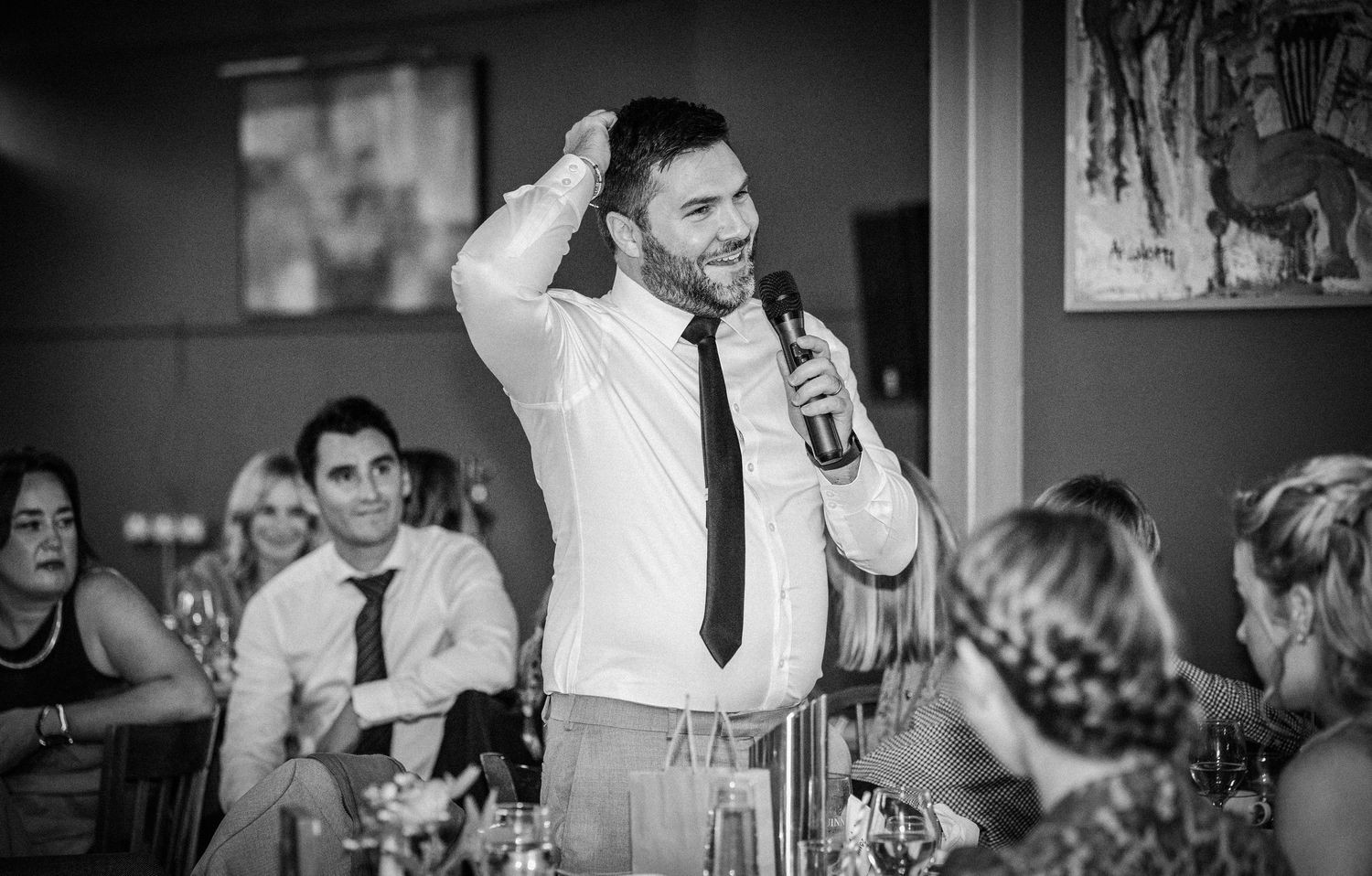 Smiling best man delivers wedding speech into microphone before seated reception guests in black and white.