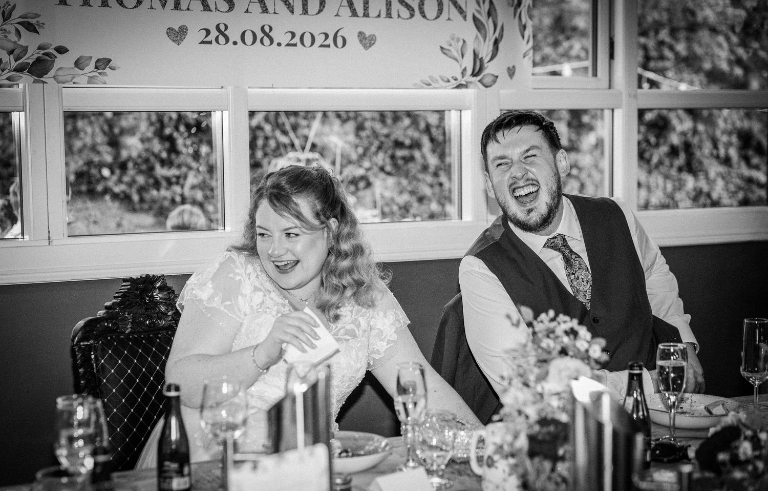 Bride and groom laughing together at the head table beneath a personalized wedding banner.