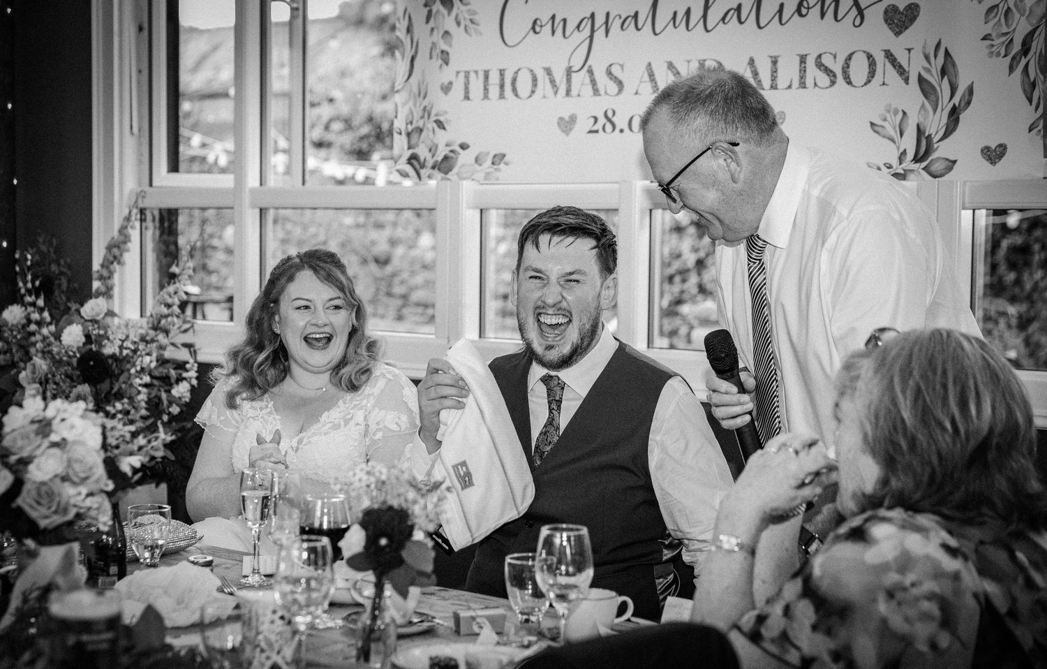 Bride and groom laughing during wedding speech at reception, congratulations banner in background.