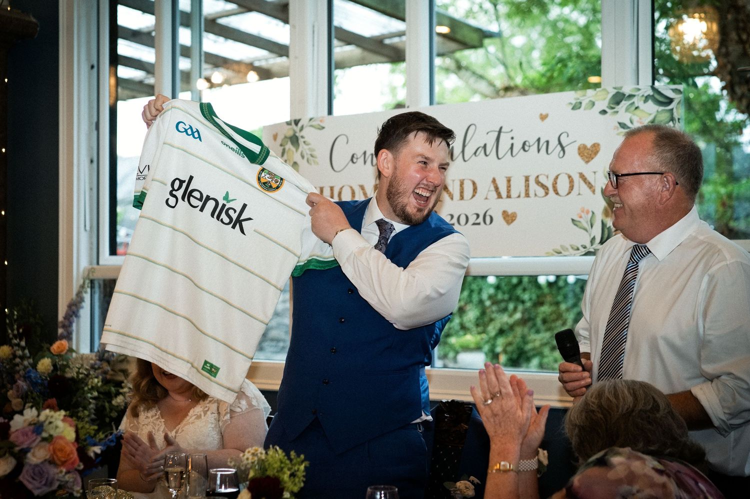 Groom in blue vest proudly holds up a GAA Offaly jersey with Glenisk sponsor at wedding reception.