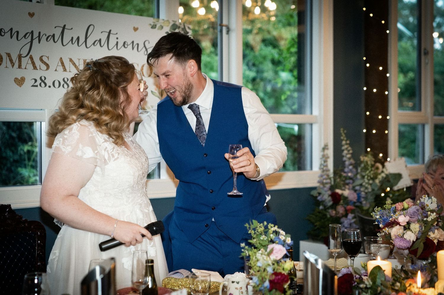 Bride and groom share a laugh during wedding reception speeches at decorated table.