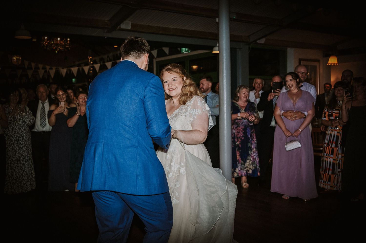 Bride and groom sharing first dance while guests clap and watch in a decorated reception venue.