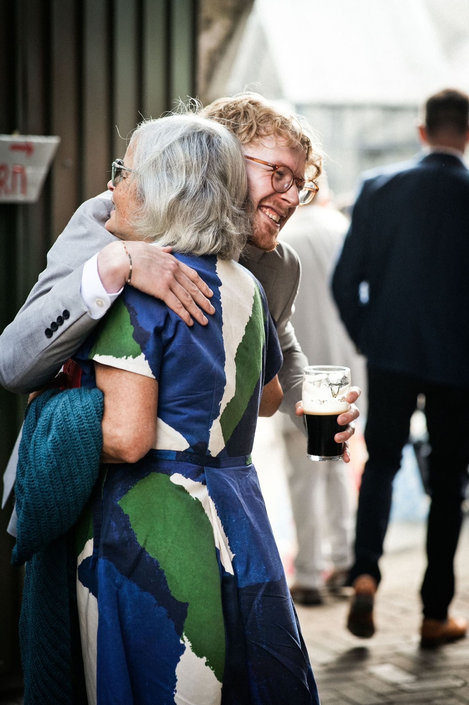 Smiling man with glasses hugging an older woman in a blue floral dress at an outdoor wedding reception.