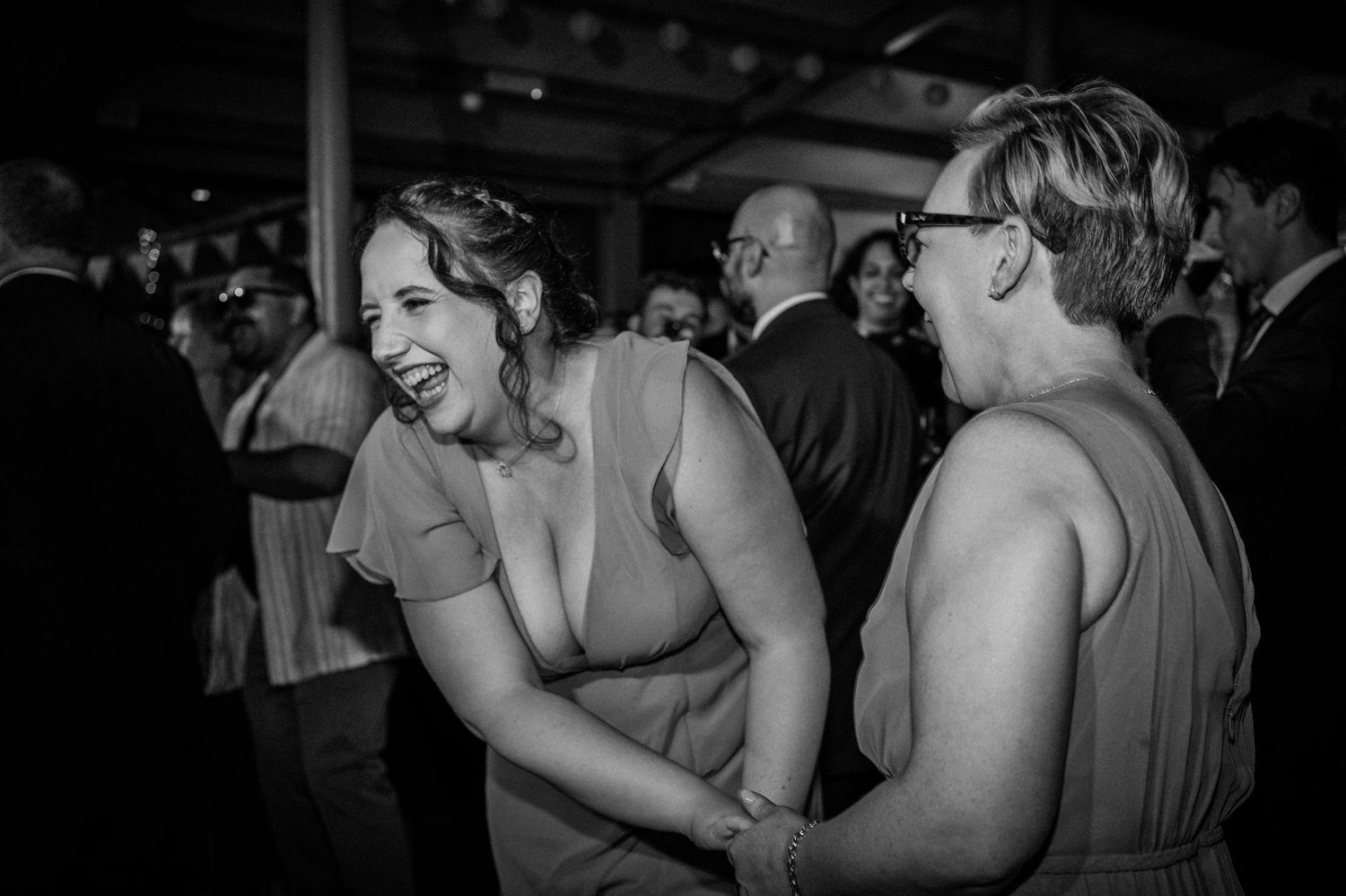 Two women in bridesmaid dresses laugh joyfully together on wedding reception dance floor in black and white.