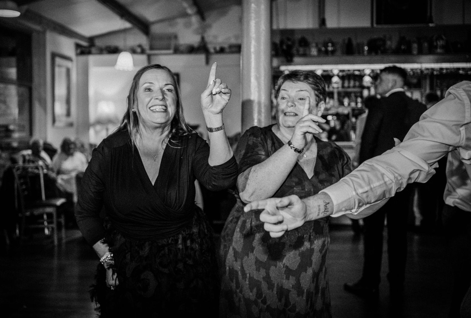 Two women dance and laugh together energetically on wedding reception dance floor in black and white photo.
