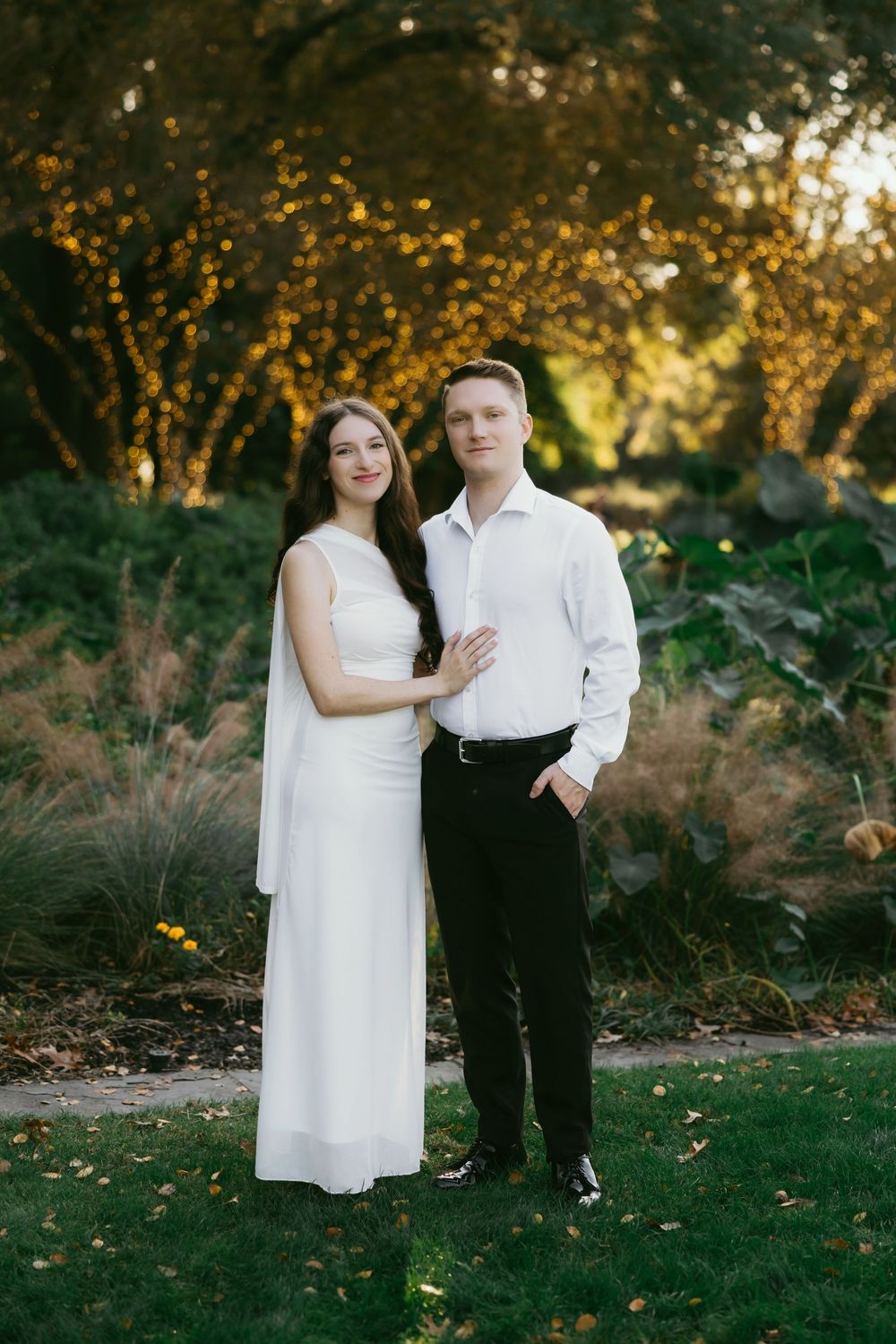 A couple poses outdoors in a garden, woman in white dress, man in white shirt and black pants.