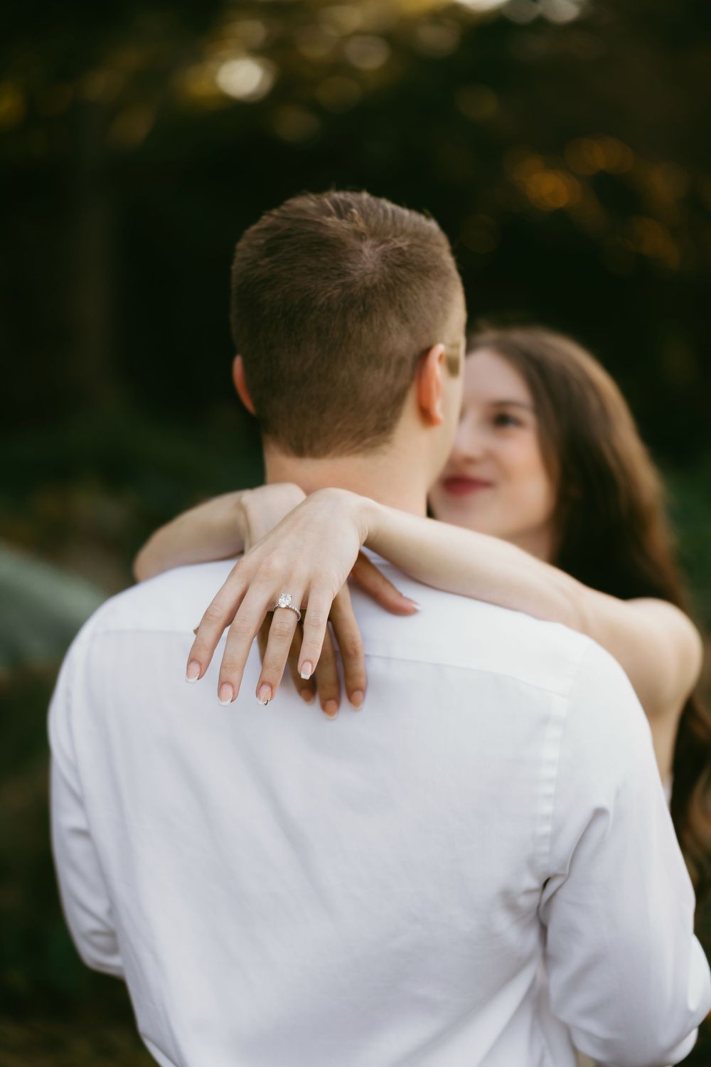 Couple embracing outdoors, woman showing diamond engagement ring on man's shoulder, romantic forest background.