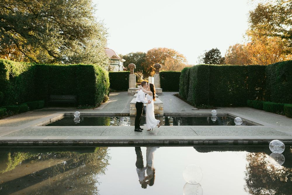 Bride and groom kissing beside a formal garden reflecting pool surrounded by manicured hedges in autumn.