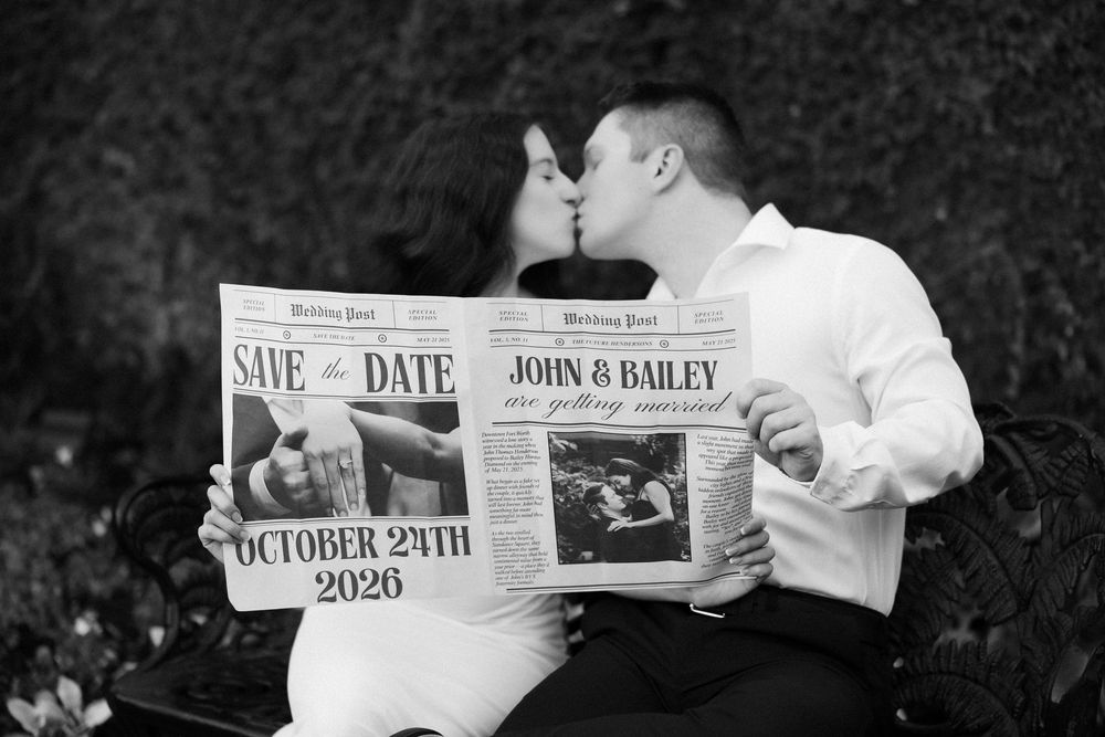 Couple kissing on bench holding newspaper Save the Date for John & Bailey, October 24th 2026.