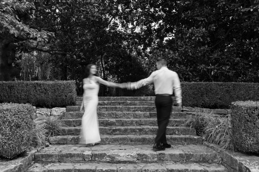 Black and white photo of a couple holding hands on stone steps surrounded by lush hedges at a garden wedding.