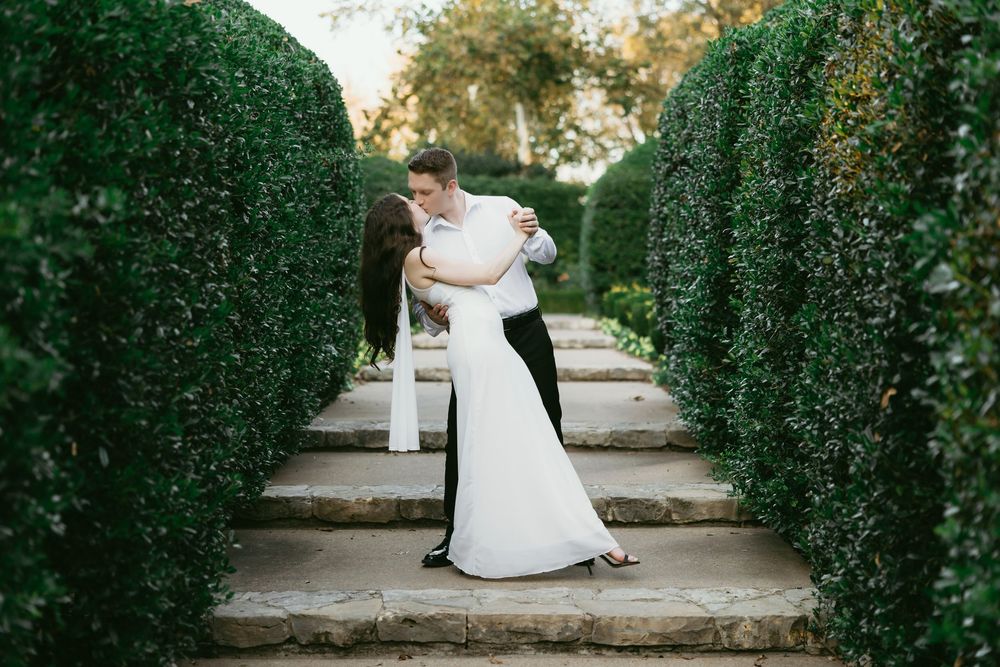 A couple shares a romantic dip kiss on stone steps surrounded by lush green hedges in a garden wedding setting.