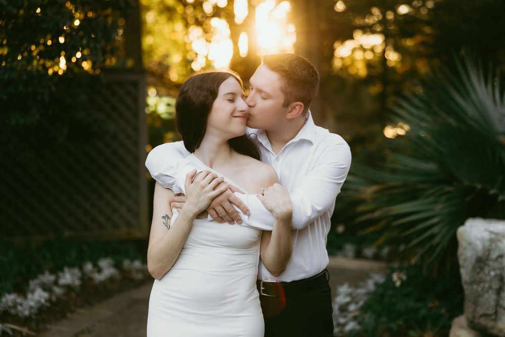 Couple sharing a romantic kiss at golden hour in a lush garden, bride in white off-shoulder gown.