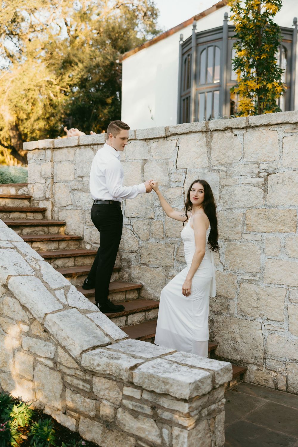 Bride in white gown holds groom's hand on stone steps outdoors during golden hour wedding photo.