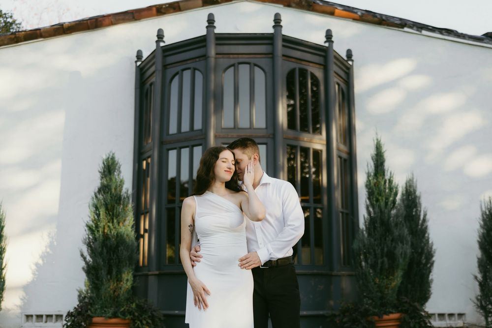 Elegant couple embracing before ornate black arched windows on white building at sunset.