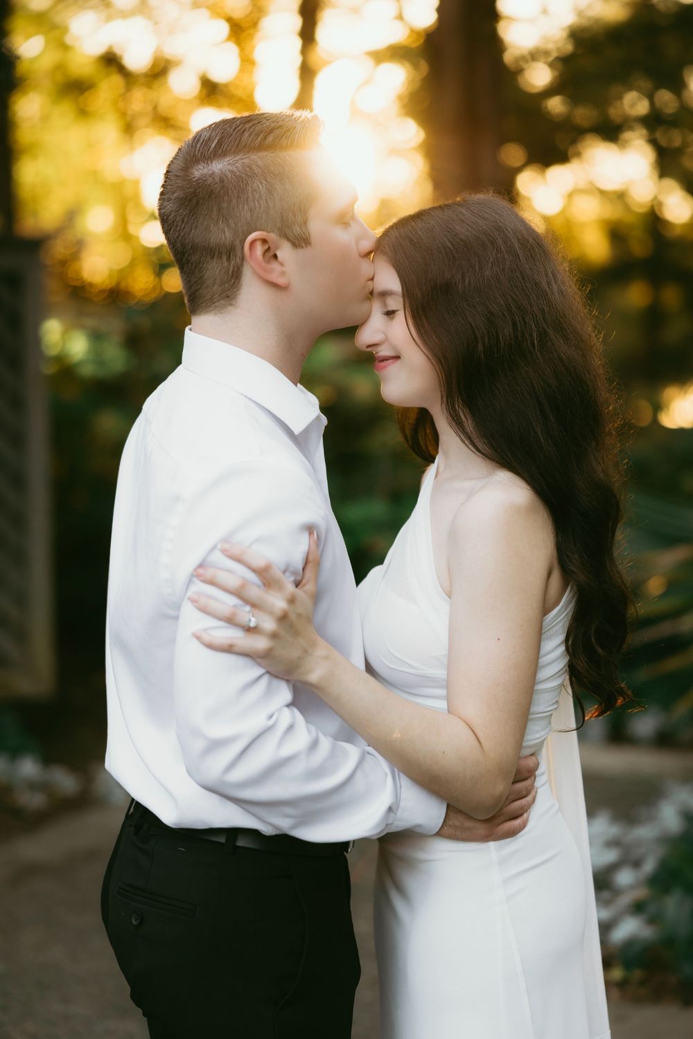 A couple shares a romantic sunset embrace outdoors, the man in white shirt kissing her forehead, woman in white dress smiling.