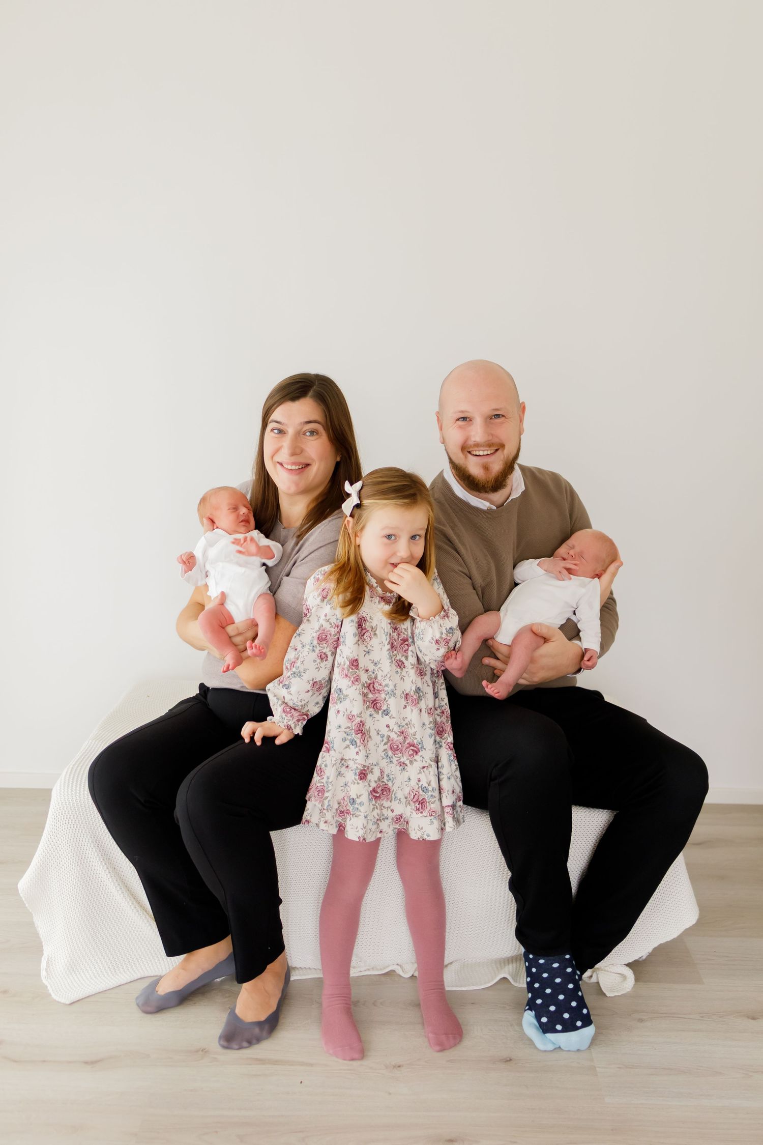 Happy family with bald father, mother, toddler girl, and twin newborns posing together on white couch.