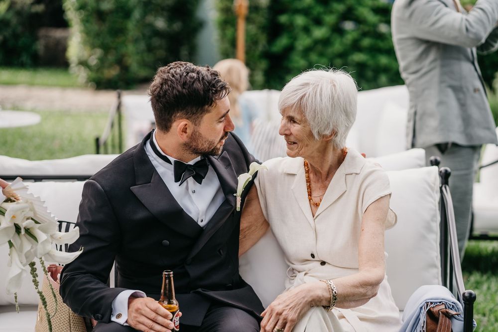 Groom in tuxedo sharing a joyful moment with elderly woman at outdoor wedding reception.