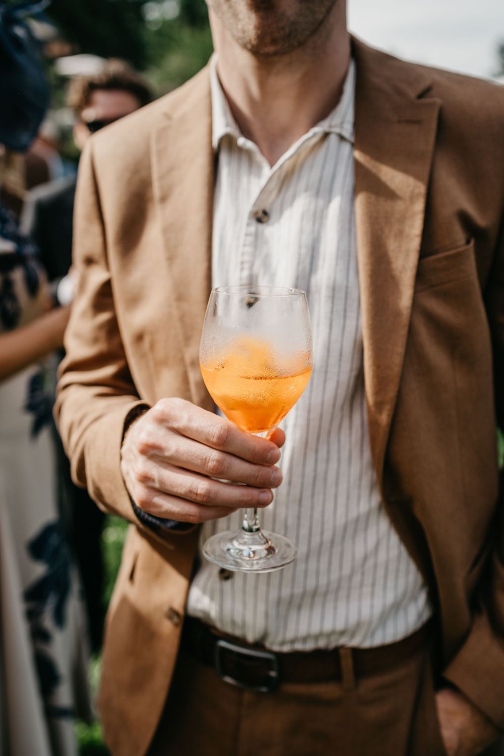 Man in tan blazer holding an orange Aperol Spritz cocktail in a wine glass at an outdoor social event.