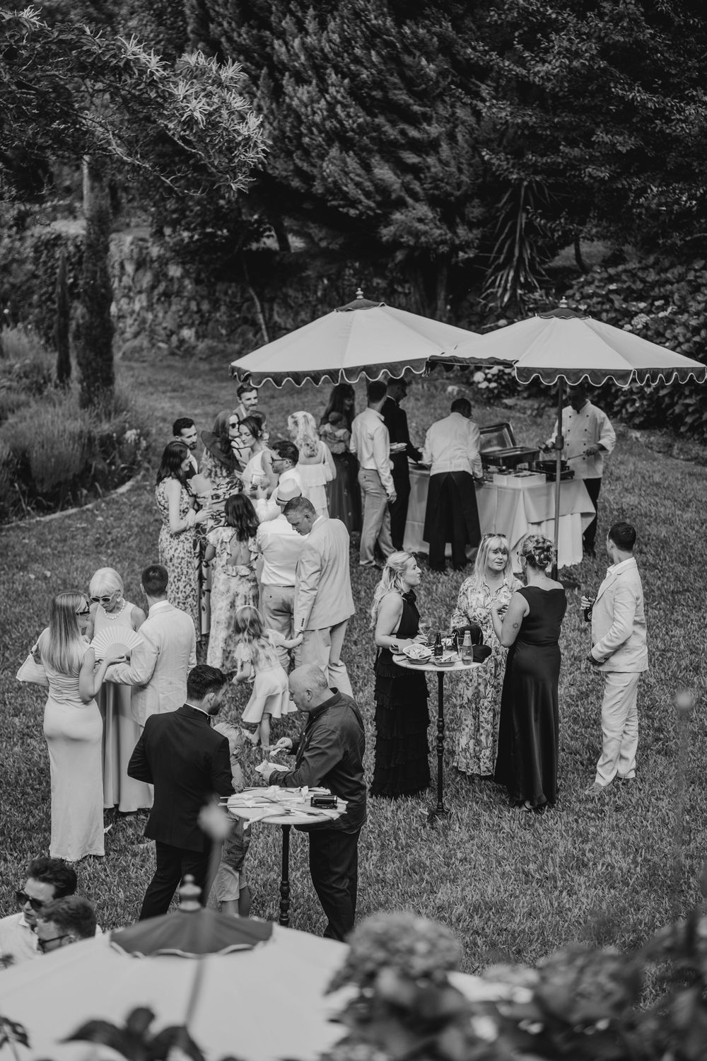 Black and white photo of elegantly dressed guests mingling outdoors under umbrellas at a garden cocktail party.