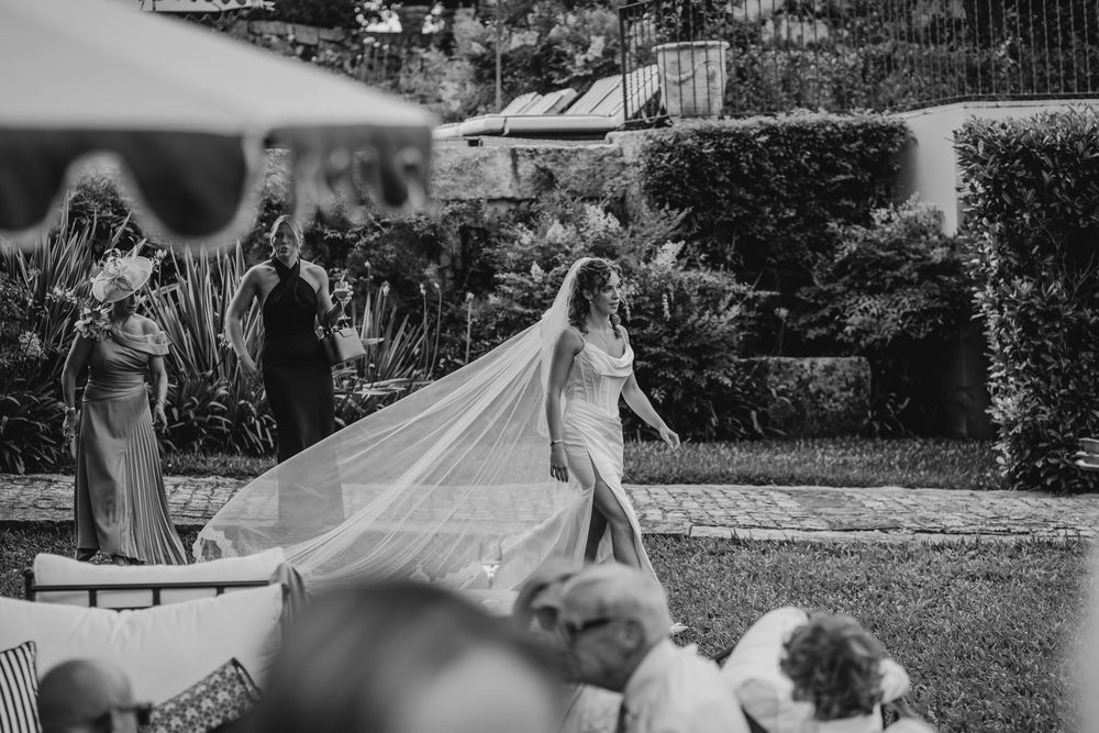 Black and white photo of a bride in a flowing gown and long veil walking through a lush garden wedding venue.
