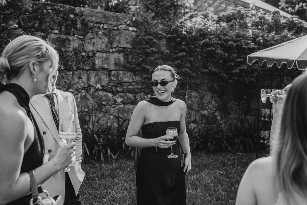 Black and white photo of women socializing outdoors at a garden party, holding champagne glasses near a stone wall.