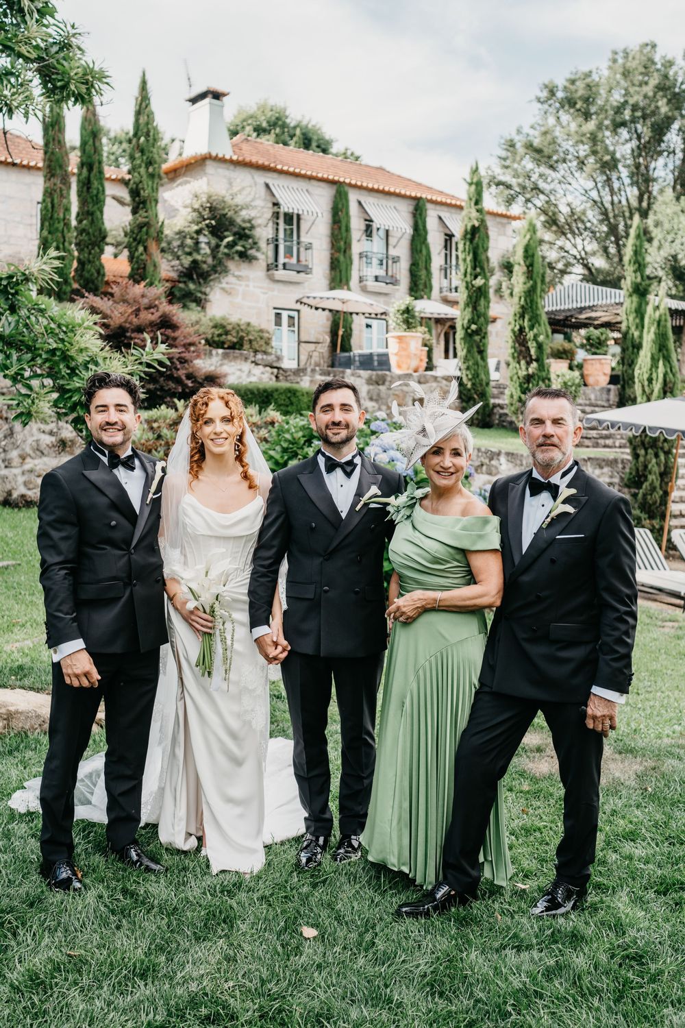 Wedding party posing outdoors at elegant stone villa, bride in white gown with veil, groom and guests in formal attire.