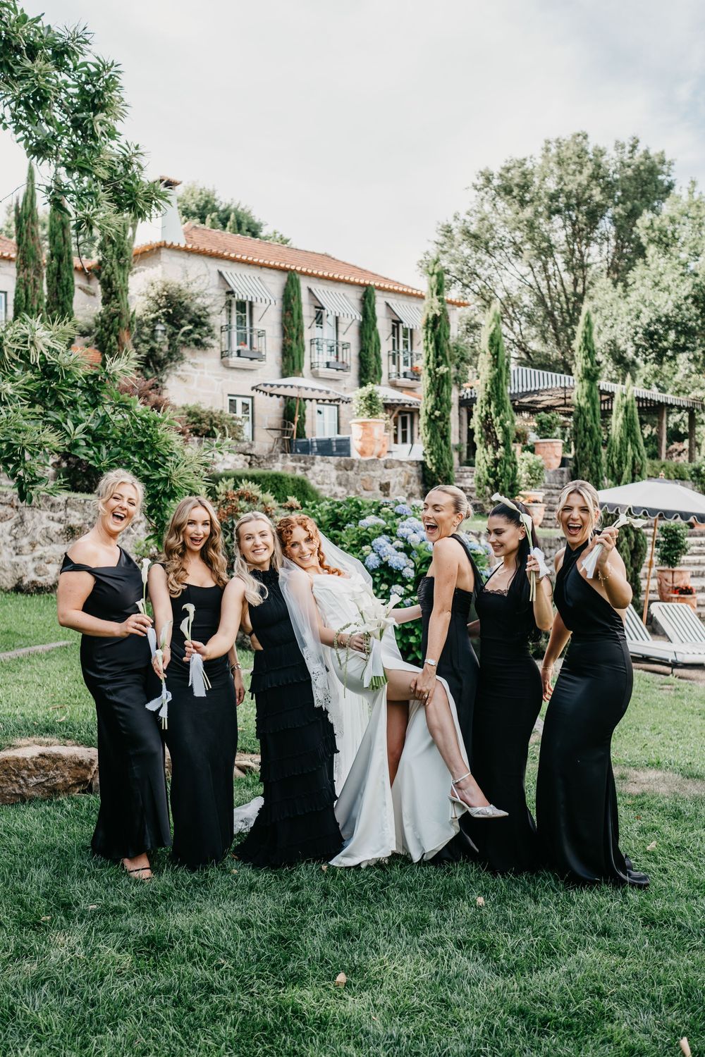 Bride in white gown poses with six bridesmaids in black dresses on green lawn before elegant stone villa.