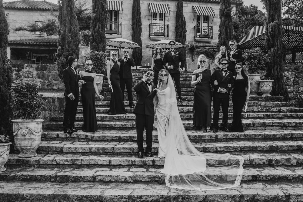 Black and white photo of a wedding party posing on stone steps outside a villa, bride and groom in front.