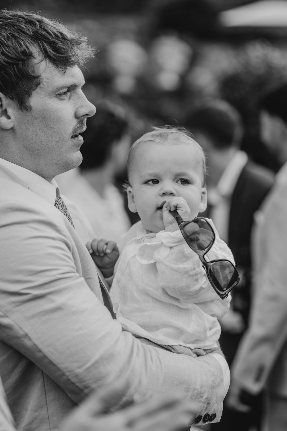Black and white photo of a man in a suit holding a baby chewing on sunglasses at an outdoor event.