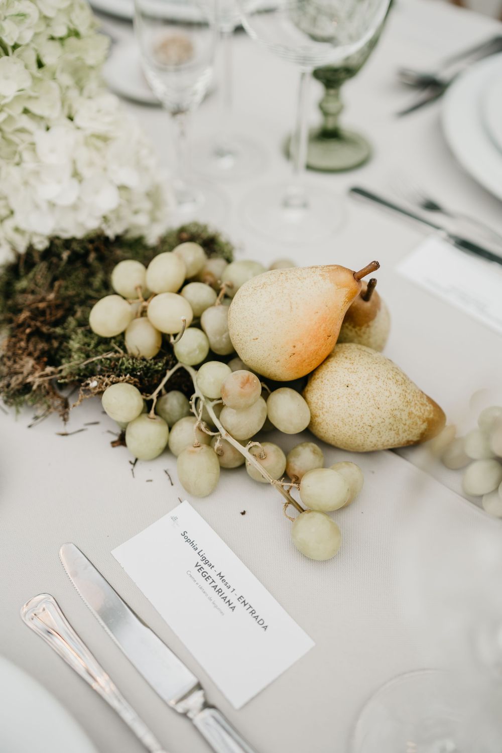 Elegant wedding table setting with green grapes, pears, white hydrangeas, moss, place card, and glassware on white linen.