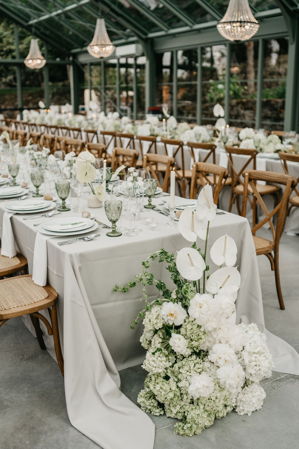 Elegant greenhouse wedding reception with white floral centerpieces, gray linen tables, cross-back chairs, and crystal chandeliers.