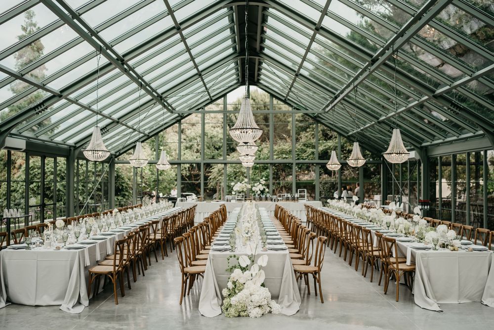 Elegant greenhouse wedding reception with long tables, floral chandeliers, white blooms, and lush greenery under a glass ceiling.