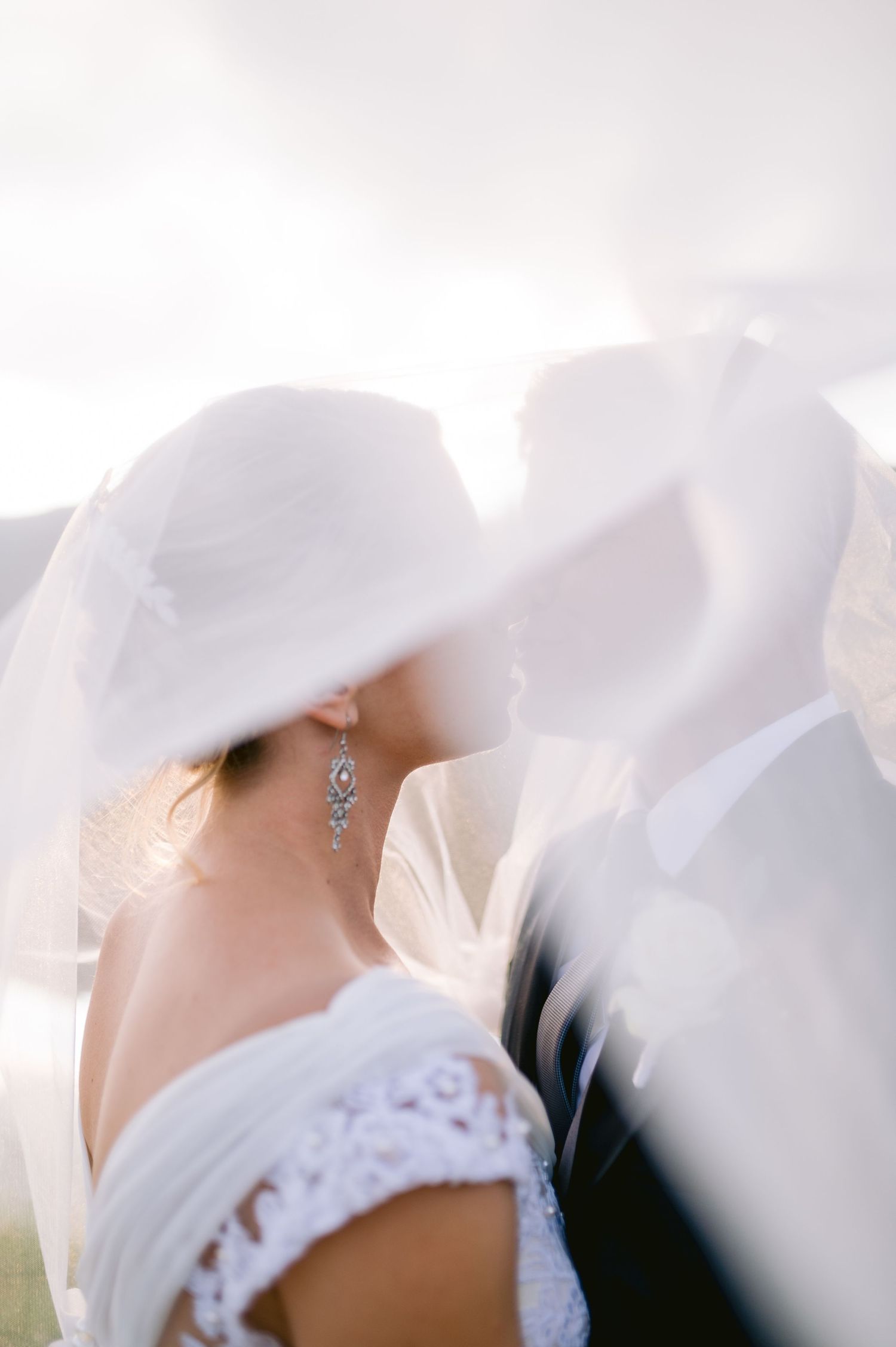 Bride and groom sharing a romantic kiss under a flowing white veil, with soft backlit glow and delicate crystal earring detail.