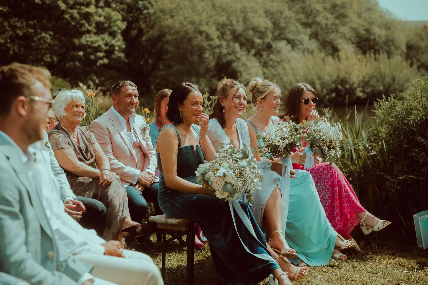 Wedding guests seated outdoors, smiling and laughing during a garden ceremony, with bridesmaids in teal dresses holding bouquets.