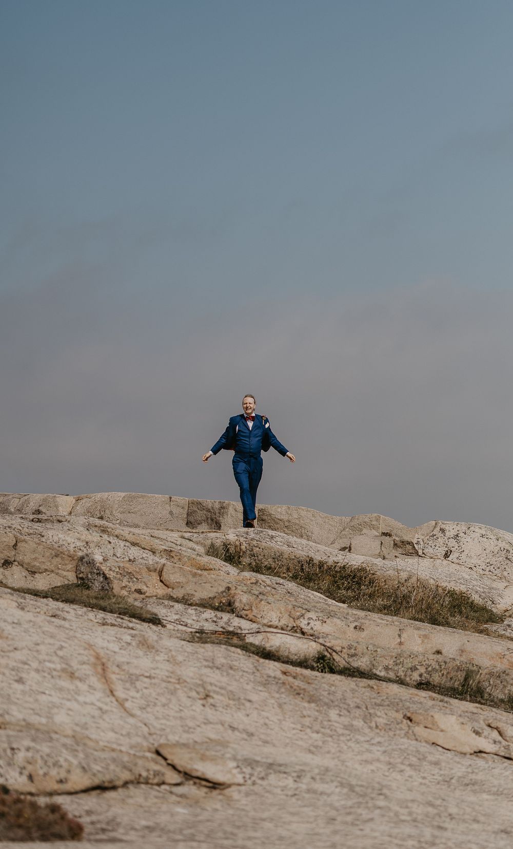A person in a blue suit walks confidently across rocky terrain under a hazy sky.