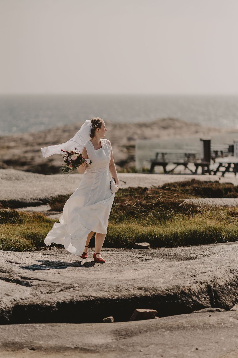 Bride in flowing white dress and veil holds bouquet on rocky coastal landscape with ocean backdrop.
