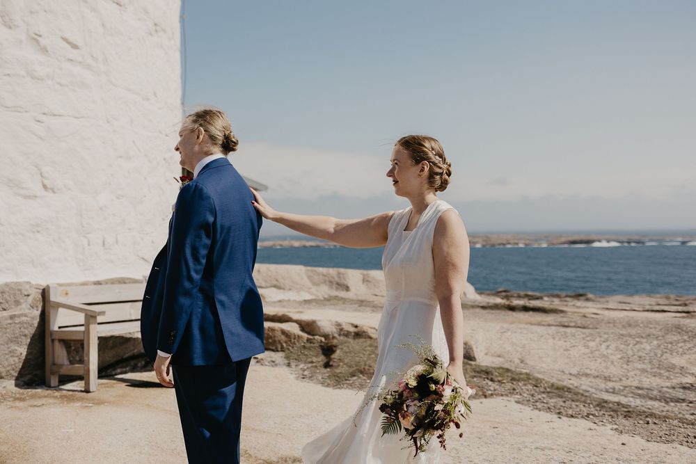 Bride taps groom on shoulder for first look moment by seaside white building on sunny wedding day.