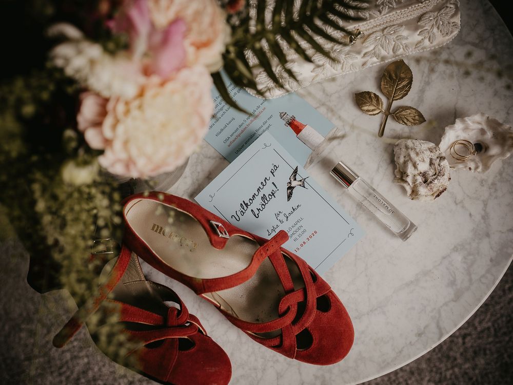 Red velvet T-strap wedding shoes beside a floral bouquet, perfume, invitation card, and seashells on marble.